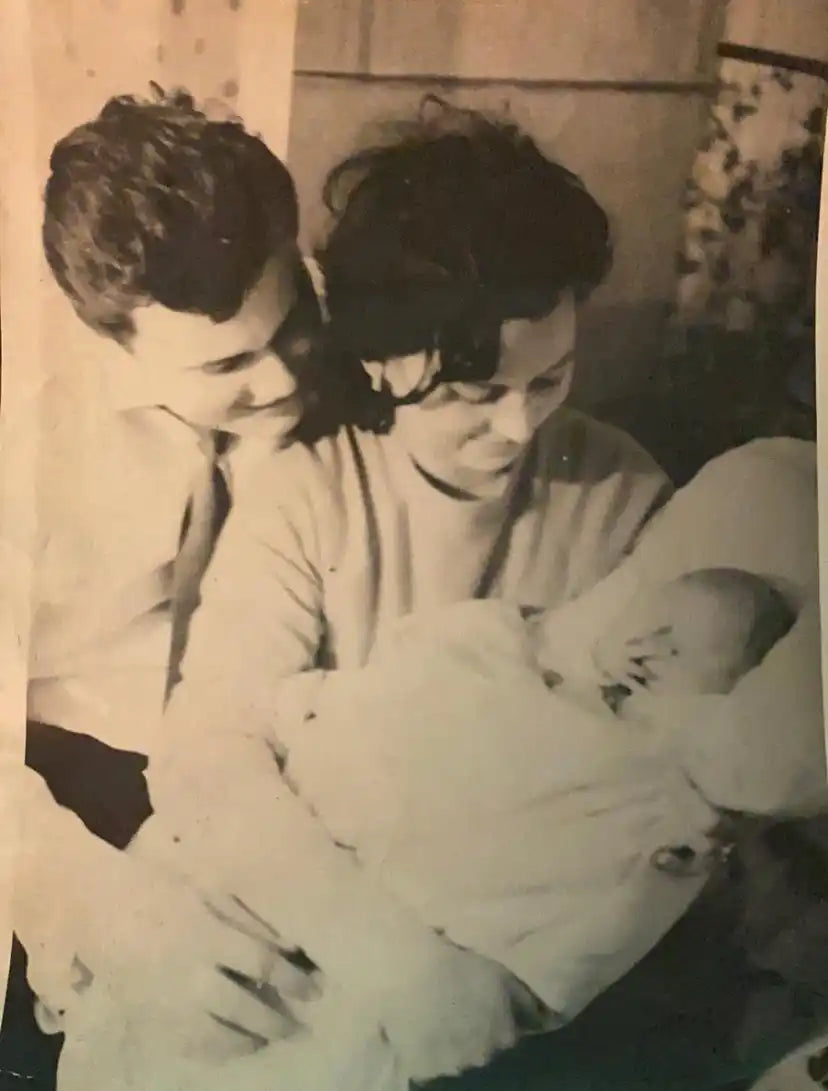 Ein Schwarzweiß-Foto eines jungen Paares, das liebevoll auf ein in eine Decke gewickeltes Baby schaut.